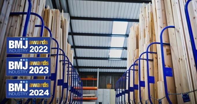 A photograph of pre-cut timber on racks inside a warehouse. On the left side, there is the three logos from the previous Builders Merchants Journal (BMJ) awards.